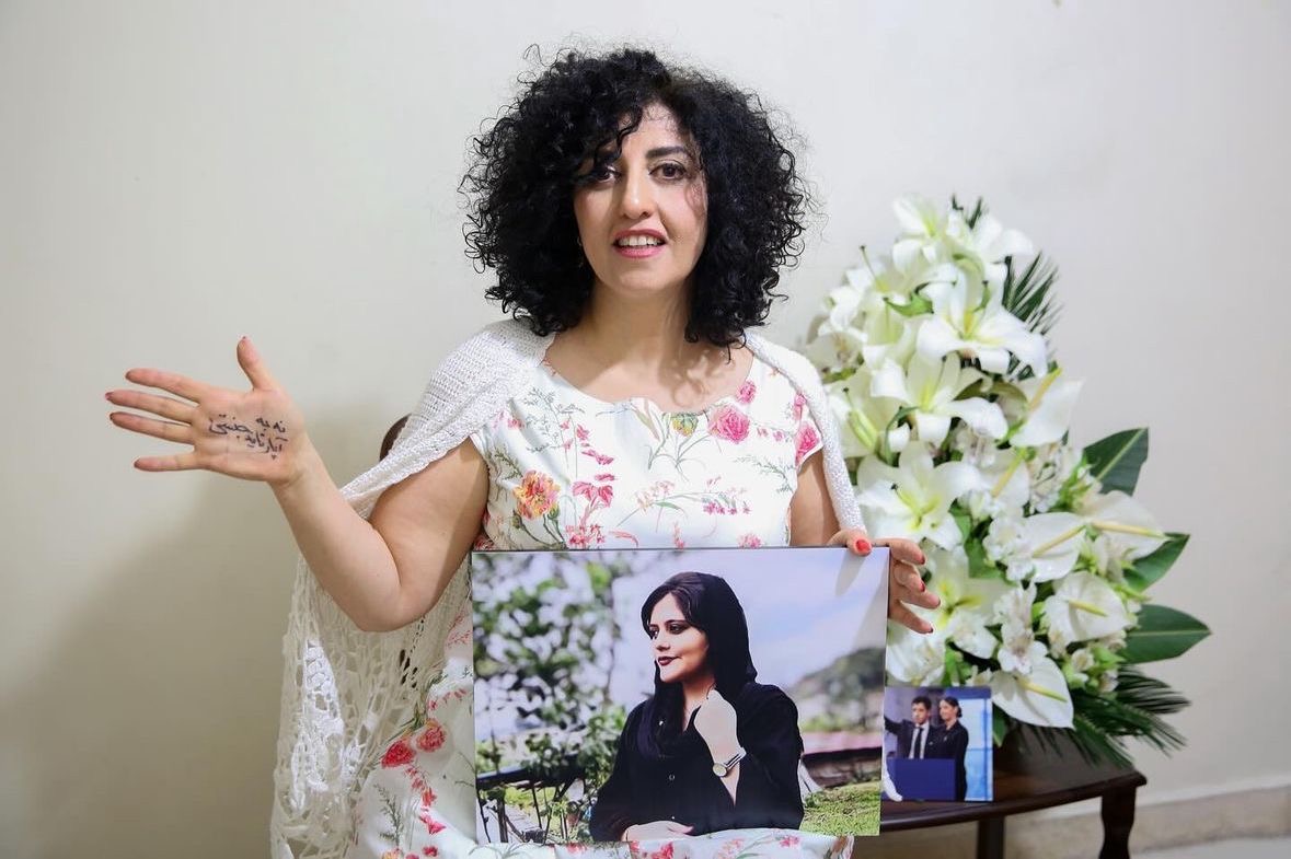 A woman with curly hair, wearing a floral dress, holds a large photo of another woman. She has writing on her palm. White lilies are in the background on a small table with a smaller framed photo.