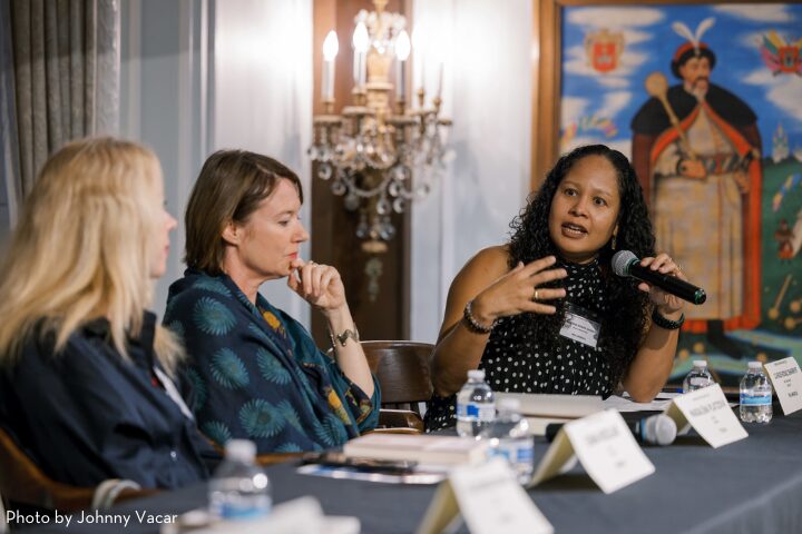 Three women are seated at a panel discussion. The woman on the right is speaking into a microphone, gesturing with her hand. A framed painting and a chandelier are in the background.