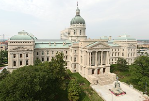Aerial view of the Indiana State Capitol building in Indianapolis. The historic structure features a large central dome, ornate details, and a symmetrical design. Surrounded by trees and a statue in the foreground.
