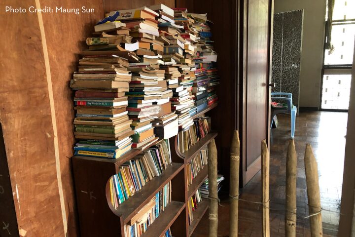 A wooden shelf overflowing with neatly arranged and stacked books in a narrow hallway. The space is filled with light from a window, casting shadows across the floor. A portion of a blue chair is visible in the adjoining room.