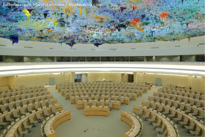 A large, modern auditorium with rows of beige seating and desks arranged in a circular pattern. The ceiling features a vibrant, colorful art installation resembling an abstract and surreal landscape.