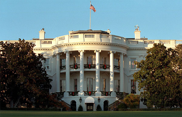 The White Houses north facade is shown with columns and a central balcony. An American flag flies atop the building. The scene is framed by green trees under a clear blue sky.
