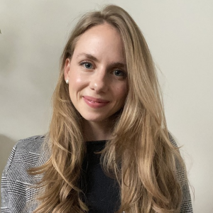 A person with long blonde hair, wearing a checkered blazer over a dark top, smiles at the camera against a plain background.