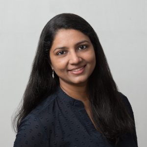 A person with long dark hair is smiling, wearing a dark, textured blouse against a light gray background.