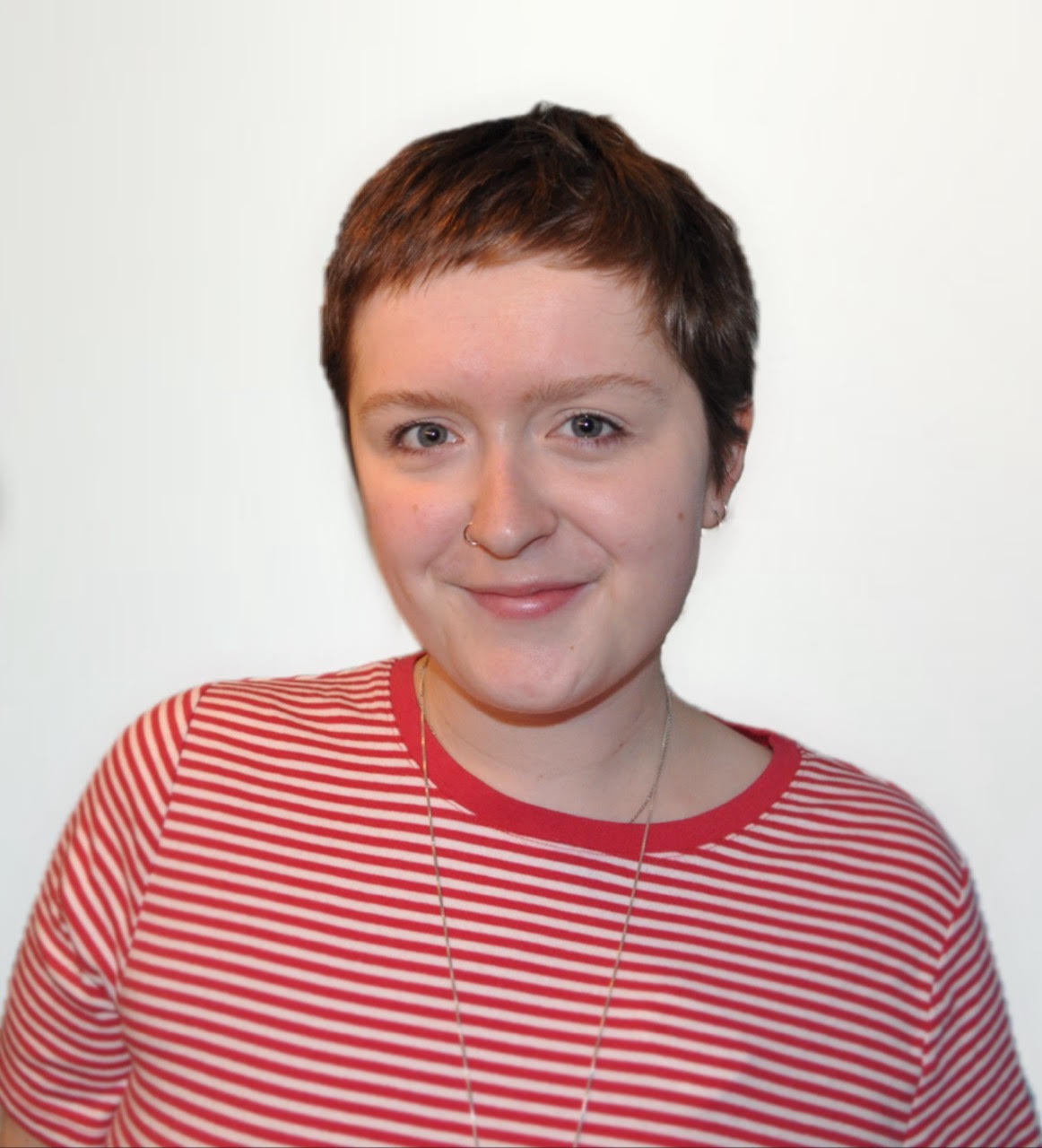 A person with short brown hair smiles at the camera. They are wearing a red and white striped shirt and a necklace. The background is plain white.