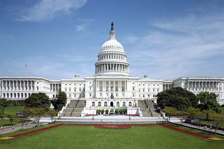 The United States Capitol Building in Washington, D.C., featuring its iconic white dome, surrounded by green lawns, trees, and a clear blue sky.