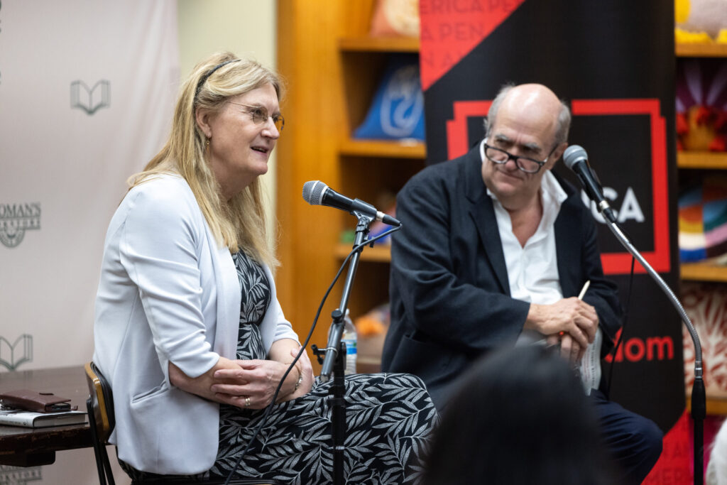 A woman and a man sit on a stage with microphones, engaged in conversation. The woman wears a light-colored jacket and patterned dress, while the man wears a black suit and glasses, holding a notebook. A banner and bookshelf are visible in the background.