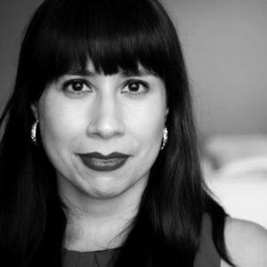 Black and white portrait of a person with long dark hair and bangs, wearing earrings. The expression is neutral, with direct eye contact. The background is blurred.