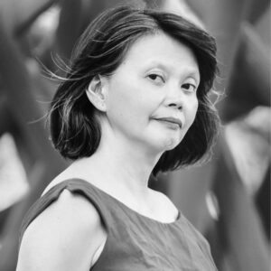 A black and white photo of a person with short, dark hair, wearing a sleeveless top. They are looking slightly off-camera with a calm expression. The background is blurred, featuring abstract shapes and patterns.