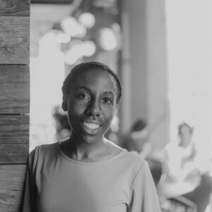 A person in a light-colored top smiles while leaning against a wooden wall. The background is softly focused, showing some lights and silhouettes of people in an outdoor setting. The image is in black and white.