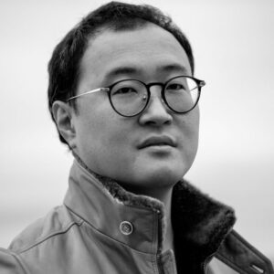 A person wearing round glasses and a jacket is looking into the camera with a neutral expression. The image is in black and white. The background is blurred.
