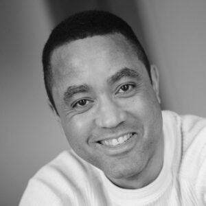Smiling person in a close-up, black and white portrait, wearing a light-colored sweater. The background is softly blurred.