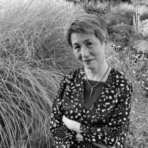 A person with short hair stands in a garden, arms crossed, wearing a patterned long-sleeve top. Tall grasses and flowers surround them. The image is in black and white.
