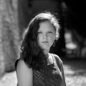 Black and white portrait of a young person with long, wavy hair and hoop earrings, wearing a sleeveless plaid top. They are standing in an alleyway with sunlight illuminating their hair. The background is slightly blurred.