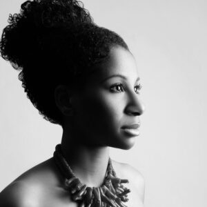 Black and white profile portrait of a person with curly hair tied up. They are wearing a chunky necklace and looking to the right. The background is a plain light color.
