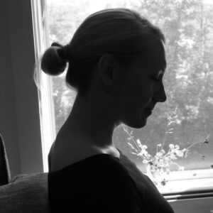 Black and white silhouette of a person with hair in a bun, sitting in profile by a window. Soft natural light highlights the serene expression. A blurred view of foliage is visible outside.