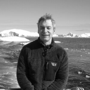 A person in a dark fleece jacket stands smiling in front of a snowy landscape and a body of water, under a clear sky.