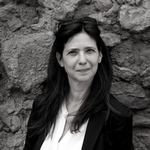 Black and white image of a woman with long dark hair, wearing a blazer and a white shirt. She stands in front of a textured stone wall, looking directly at the camera.