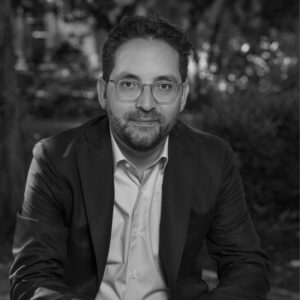 A man with short hair, beard, and glasses is sitting outdoors, wearing a suit jacket and light-colored shirt, with a neutral expression. The background is blurred greenery. The image is in black and white.