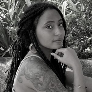 Woman with long, dark, braided hair and a tattoo on her shoulder, sitting sideways outdoors. She rests her chin on her hand and looks towards the camera. Background features lush green foliage. Image is in black and white.