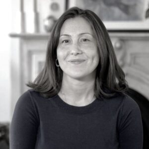 A person with shoulder-length hair is smiling slightly while wearing a dark top. They are in a room with a fireplace and framed artwork in the background. The image is in black and white.
