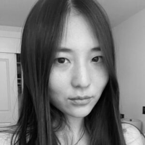 Black and white portrait of a person with long, straight hair, facing the camera. The background shows a partially visible door and wall. The expression is neutral, and the lighting is soft.