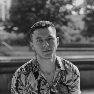 A person with short hair wearing a patterned shirt looks at the camera. The background is outdoors, featuring greenery and blurred structures. The image is in black and white.