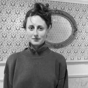 A person with short hair in a high bun stands in front of a patterned wallpaper with an oval mirror. They are wearing a high-neck sweater and looking calmly at the camera. The image is in black and white.