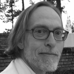 Man with glasses and short hair, wearing a light-colored shirt, looking at the camera. He has a beard and is outdoors, with trees and a brick wall in the background. The image is in black and white.