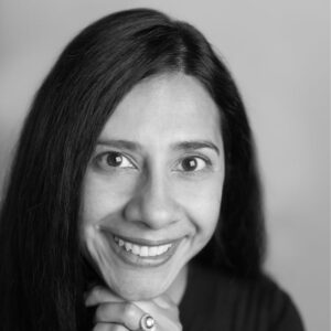 Black and white portrait of a person with long dark hair, smiling while resting their chin on their hands. The background is smooth and out of focus, highlighting the subjects face.