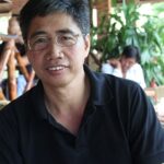 A man wearing glasses and a black polo shirt smiles at the camera. The background features blurred greenery and a person seated at a table.