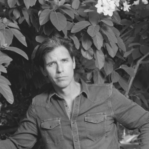 A person is sitting in front of large leafy branches with white flowers. They are wearing a denim shirt and looking directly at the camera. The image is in black and white.