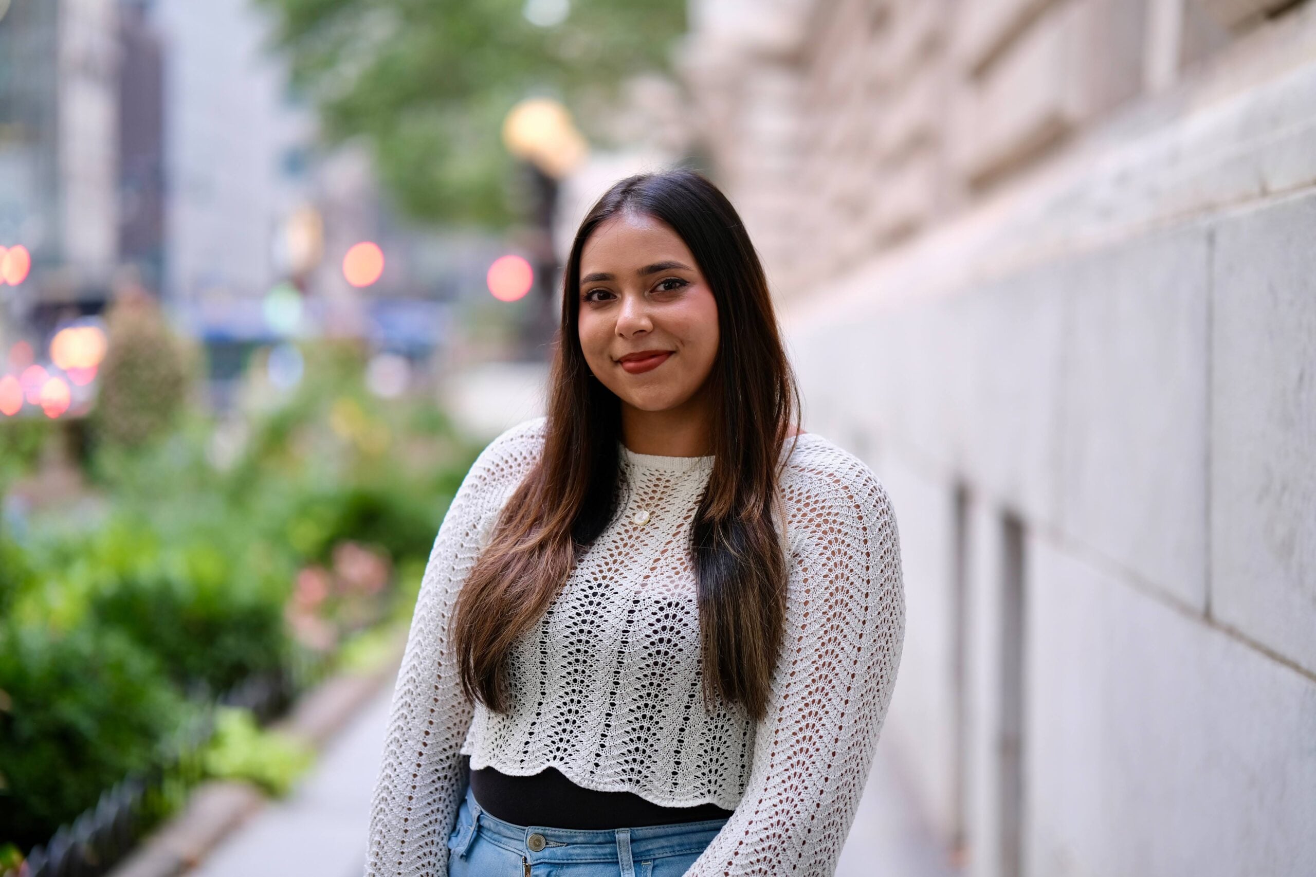 A woman with long dark hair stands outdoors, wearing a white knit top and jeans. She is smiling with a blurred cityscape and greenery in the background.