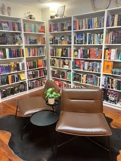A cozy reading nook with two brown leather chairs and a small round black table. The table holds a potted plant. The backdrop features white bookshelves filled with colorful books and decorations. The floor has a dark rug.