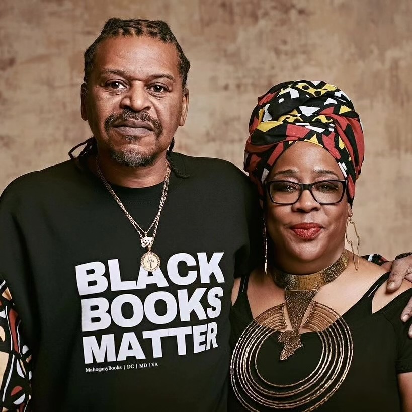A man and a woman stand together, smiling. The man wears a Black Books Matter shirt and has braided hair. The woman wears a colorful headwrap and jewelry. The background is a textured tan surface.