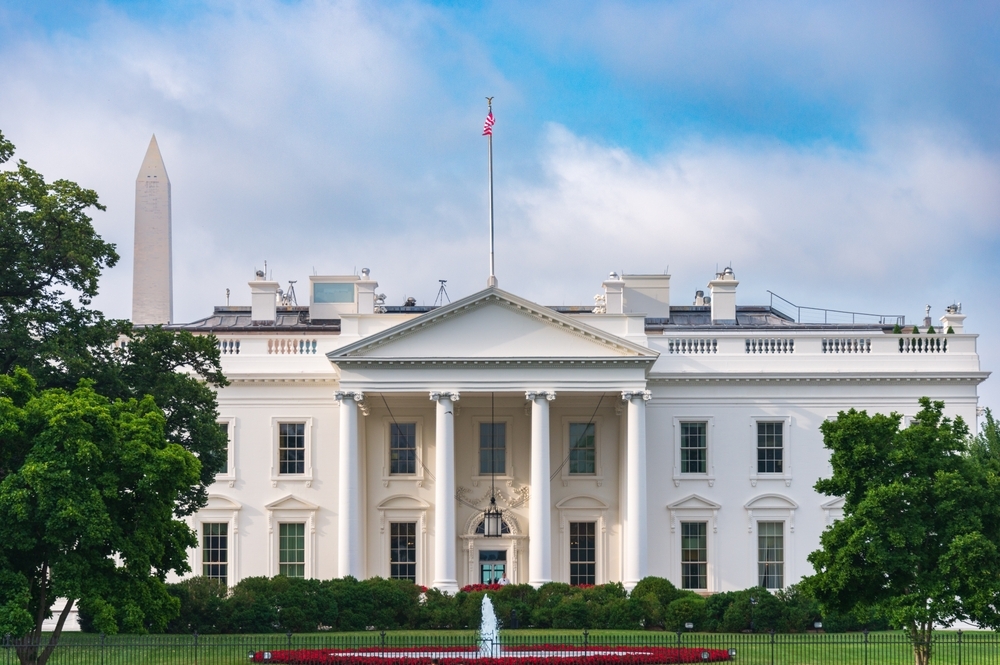 The image shows the White House with its iconic white columns and portico. The American flag flies atop the building. The Washington Monument is visible in the background, and trees frame the scene. A small fountain is in front of the building.