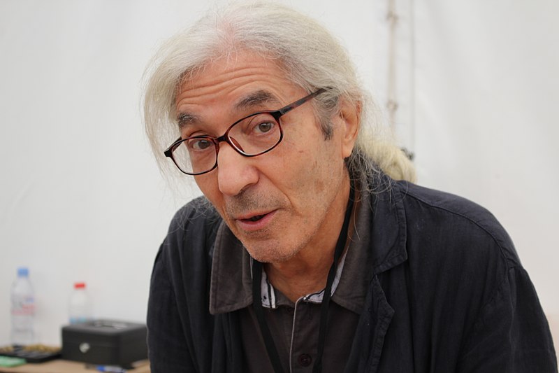 An older man with long gray hair and glasses stands indoors. He is wearing a black jacket over a gray shirt and is looking directly at the camera. The background is plain white.