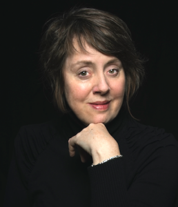 A woman with short brown hair, known among world literature speakers, poses in a black turtleneck against a dark background. Smiling slightly, she rests her chin on her hand, showcasing the bracelet on her wrist.