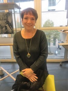 A woman with short hair smiles, standing indoors. She wears a dark long-sleeve top, a long necklace, and a bracelet. Behind her, large windows reveal trees and a building—a serene backdrop for one of the leading world literature speakers.