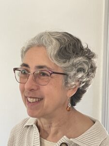 A person with short gray curly hair and glasses smiles while looking to the side, exuding the wisdom of world literature speakers. They are wearing a light-colored top and dangling earrings, set against a plain, light-colored wall.