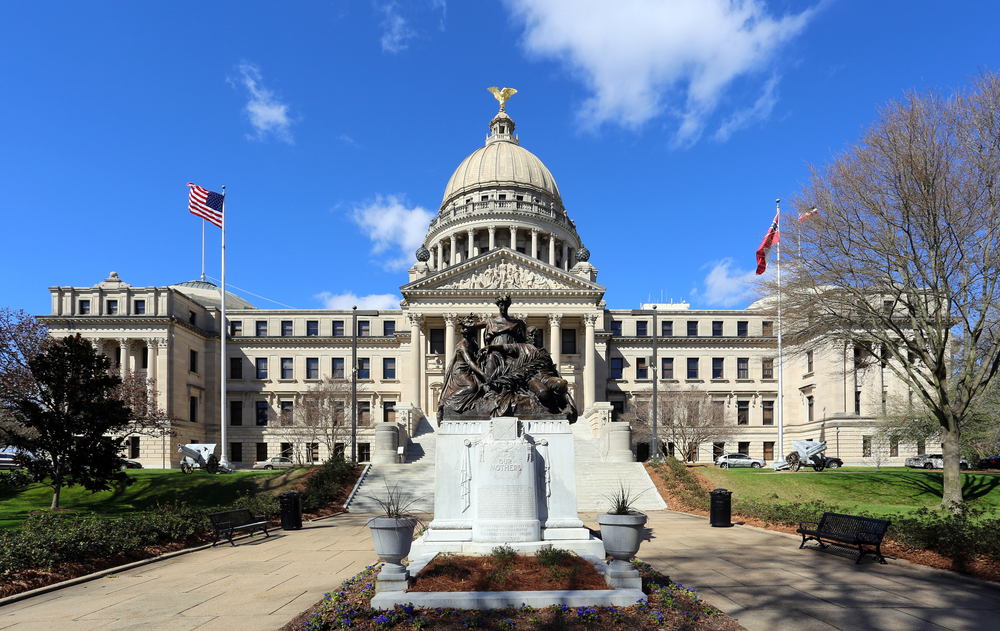 The image shows a large, historic building with a prominent dome, topped by a golden eagle statue. A statue is situated in front, with American and state flags on either side. Lush greenery surrounds the building under a clear blue sky.