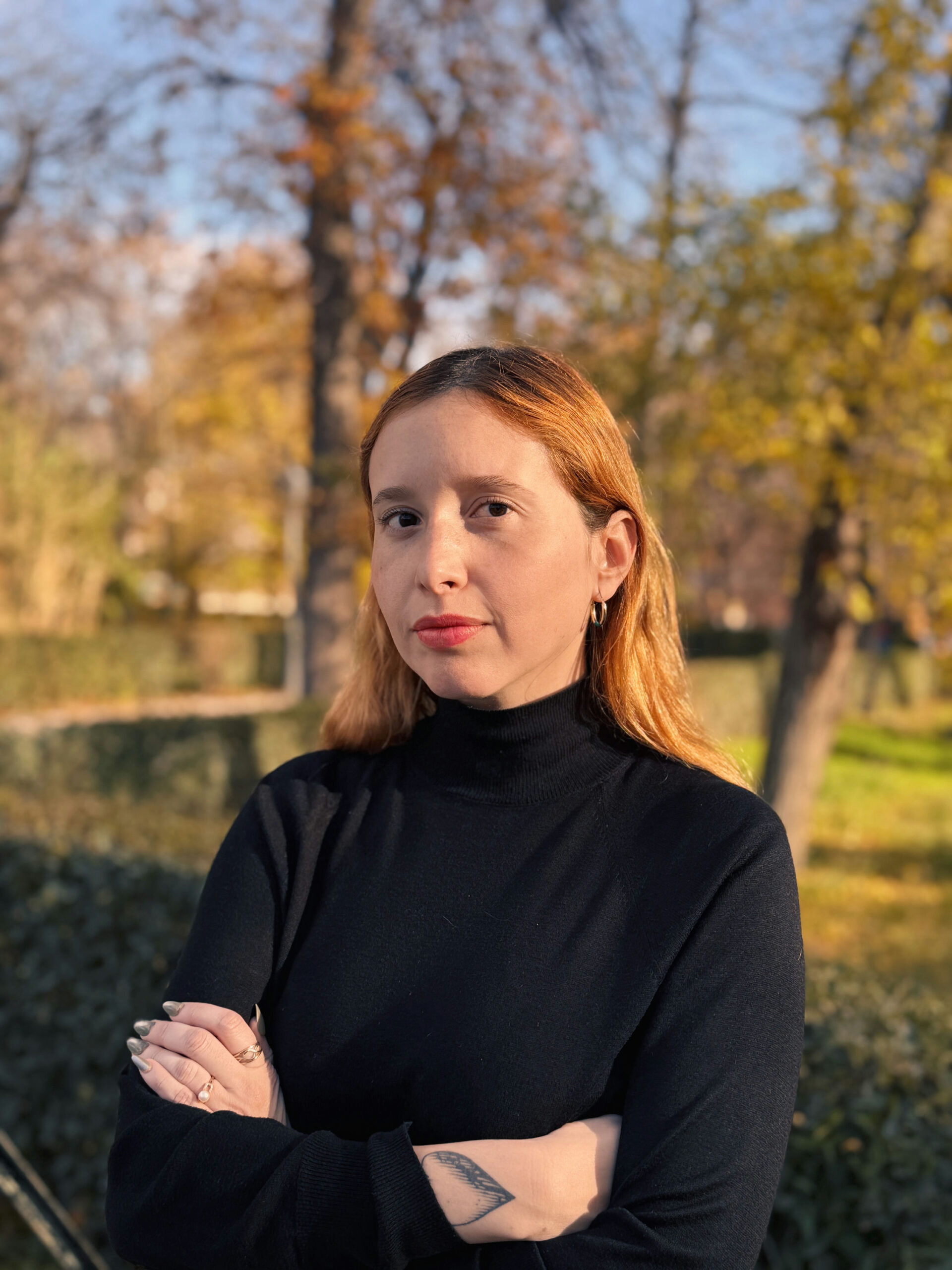 A person with long hair stands outdoors with arms crossed, wearing a black turtleneck. The background is a park with trees in autumn colors, featuring green, yellow, and brown leaves. The lighting is bright, suggesting a sunny day.