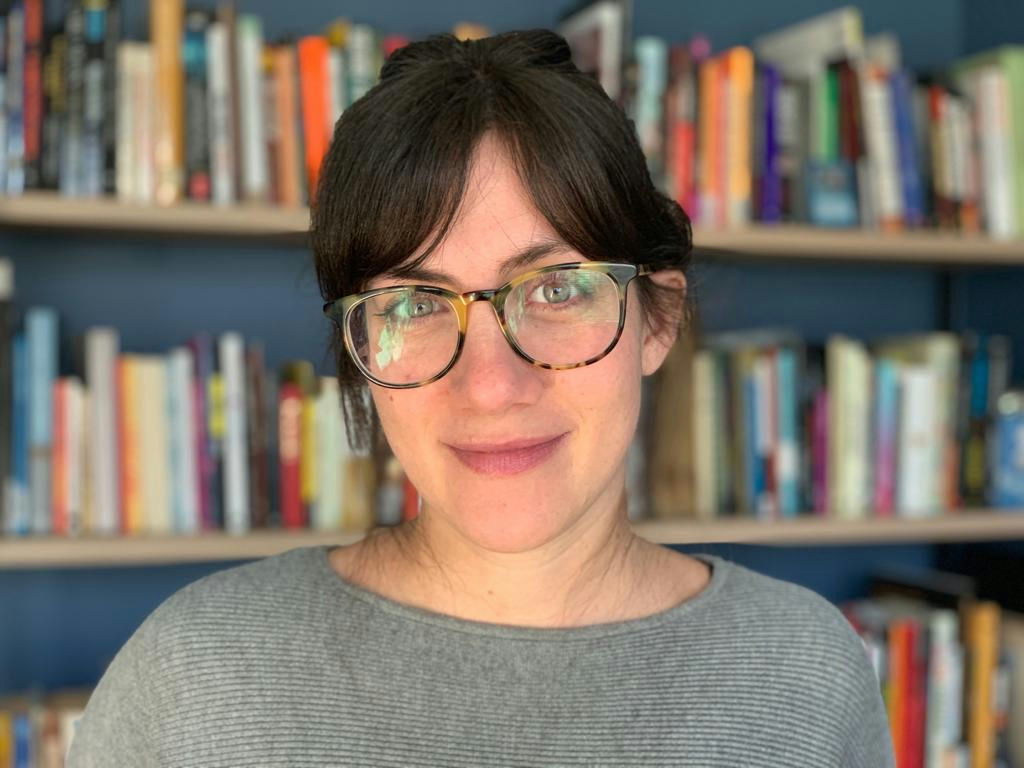 A person with glasses and long dark hair smiles softly while standing in front of a bookshelf filled with books. They are wearing a gray sweater, and the background shows colorful book spines.