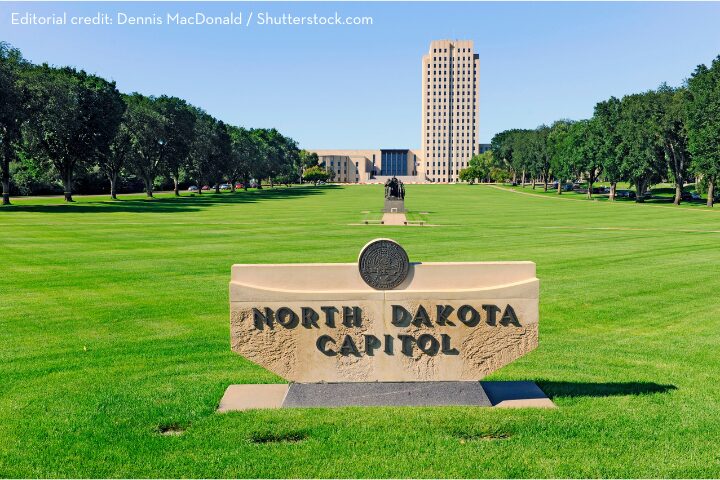 The image shows the North Dakota State Capitol building, a tall structure with rectangular windows, set against a clear blue sky. In the foreground, a sign reads North Dakota Capitol surrounded by well-manicured lawns and trees.