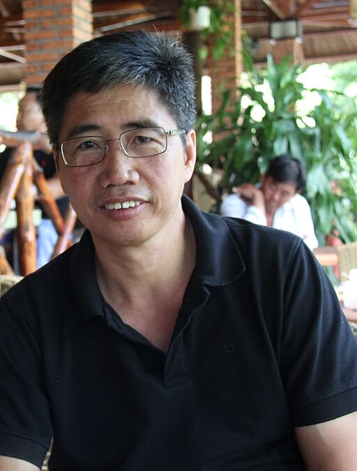 A man wearing glasses and a black polo shirt is smiling slightly, seated in an outdoor café. In the background, there are people sitting and greenery visible.