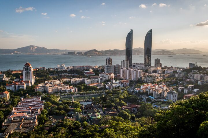 Aerial view of a coastal cityscape with modern skyscrapers, including two tall, curved buildings. The city is surrounded by lush greenery and water, with mountains visible in the distance under a clear sky.