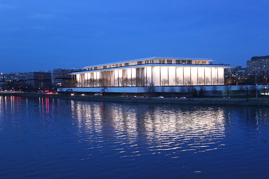 A modern building with large windows is illuminated against a dusk sky, reflecting in the calm river in the foreground. Surrounding the structure are other urban buildings, with trees lining the riverbank.