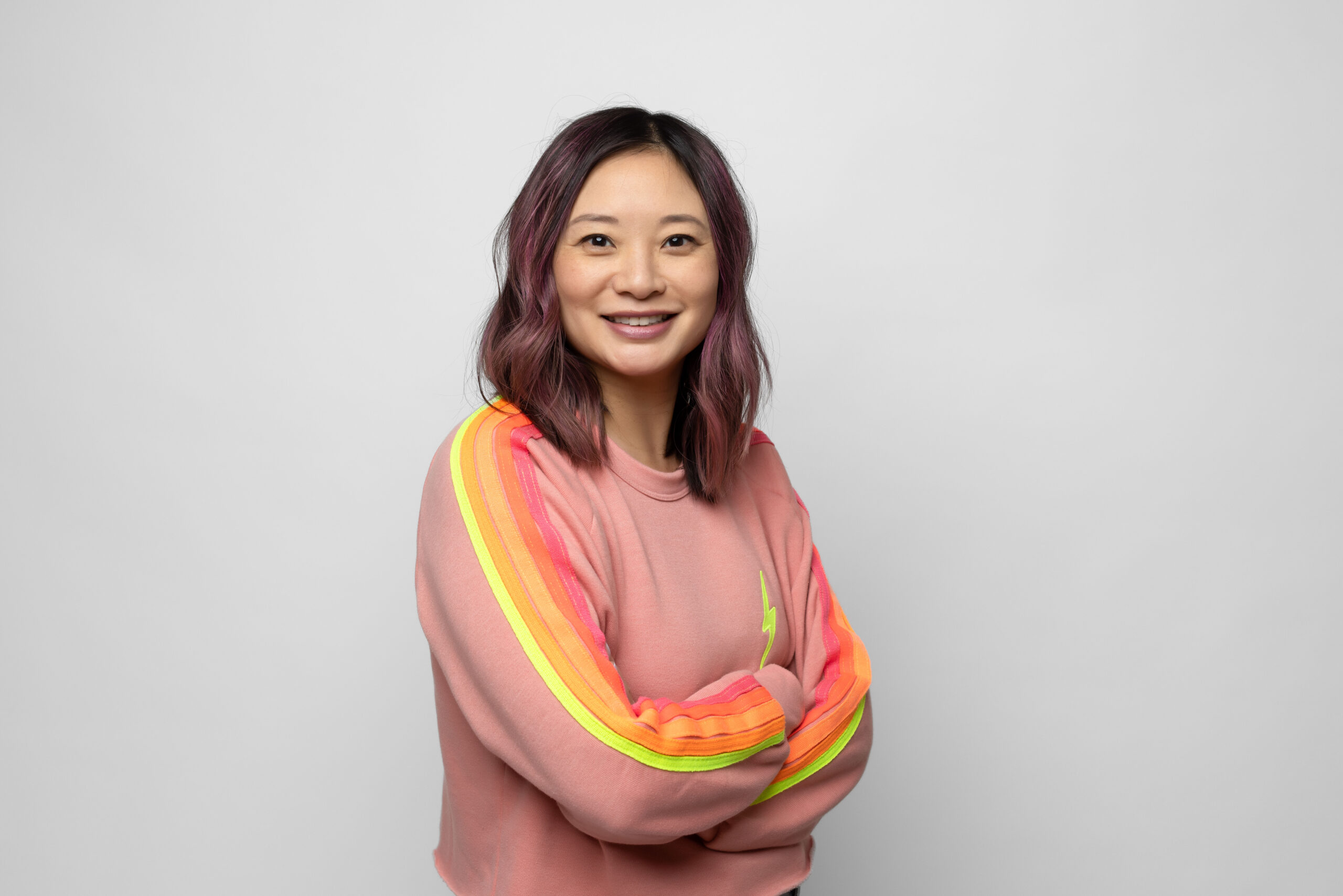 A person with shoulder-length hair, smiling, wearing a pink sweatshirt with yellow and orange striped sleeves, stands against a plain grey background. Arms are crossed casually.