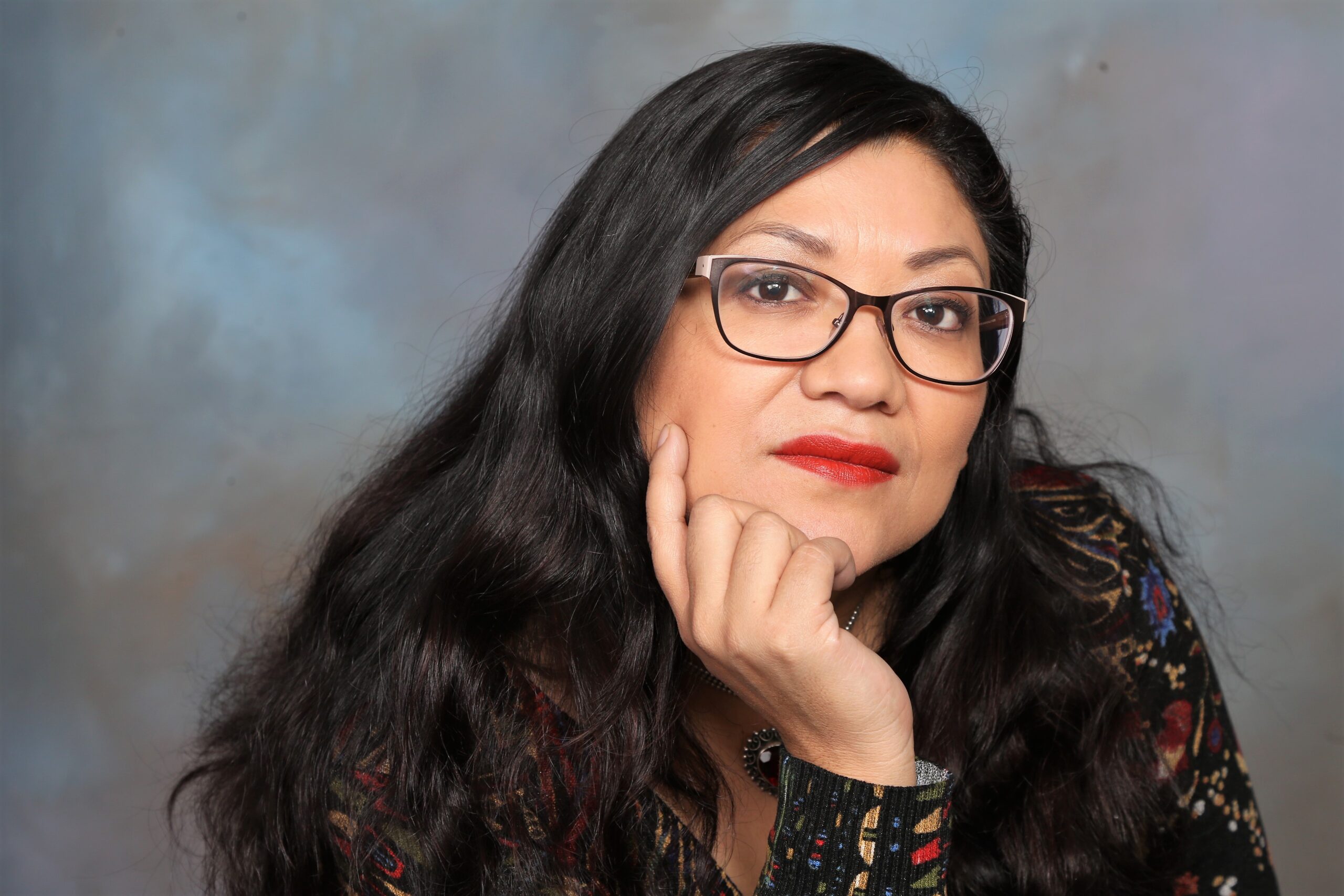 A person with long dark hair and glasses rests their chin on their hand, gazing forward. They are wearing a patterned top, seated against a blurred, muted background.