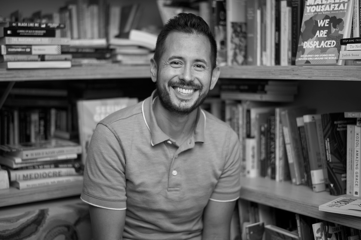 A person with short hair and a beard smiles while sitting in front of a bookshelf filled with various books. They are wearing a short-sleeved polo shirt and are surrounded by shelves of books. The image is in black and white.